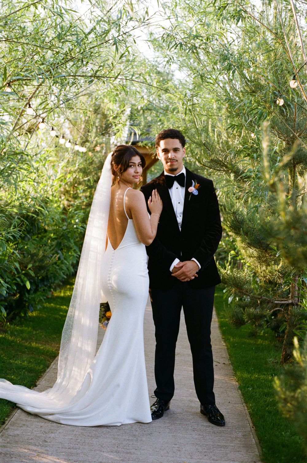 A bride and groom pose together on a garden path surrounded by lush greenery.  Shot on 35mm film.
