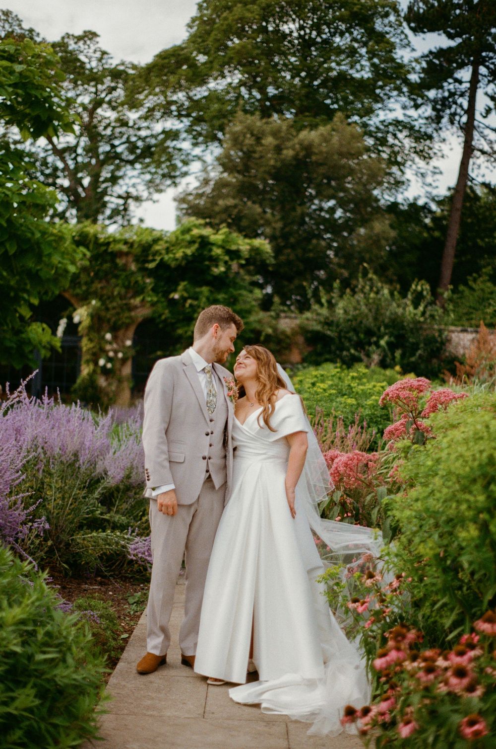 A couple shares romantic moments in a lush garden pathway surrounded by vibrant purple and pink blooming flowers.