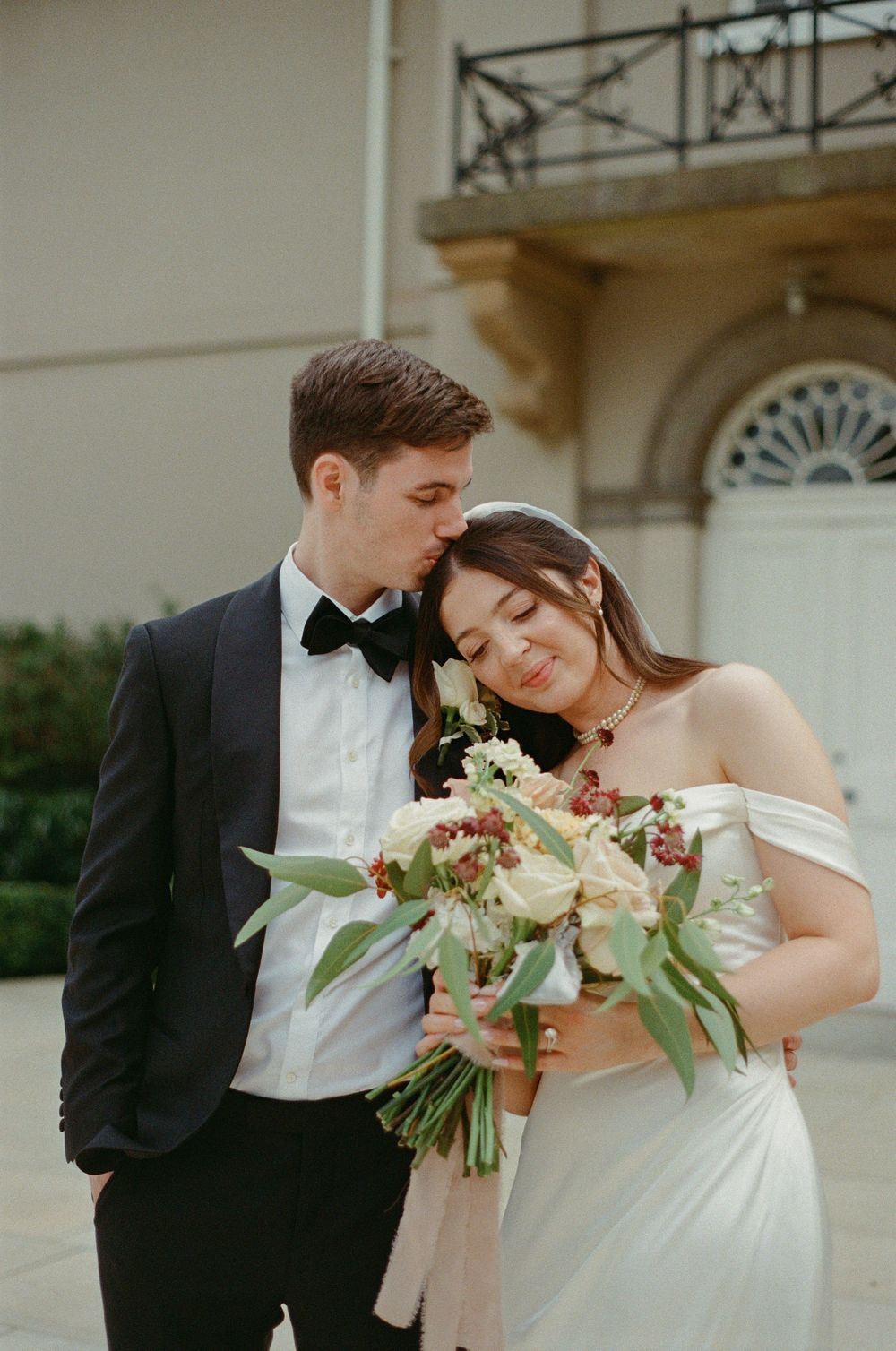A romantic wedding couple shares an intimate moment while holding a beautiful bouquet of white flowers and greenery.