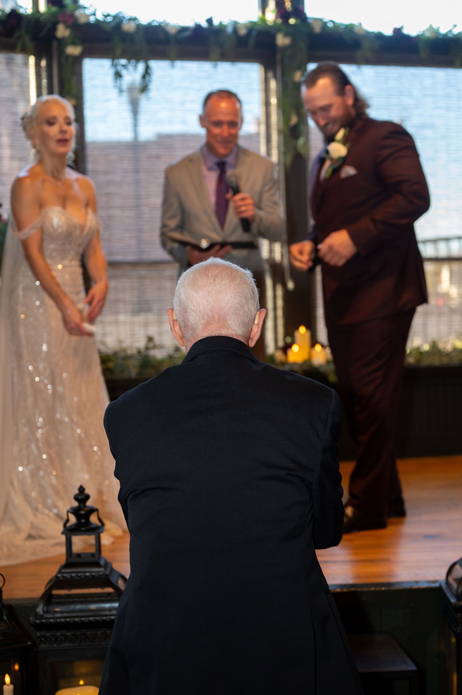 A wedding ceremony scene with people gathered on a stage near a waterfront view at sunset.