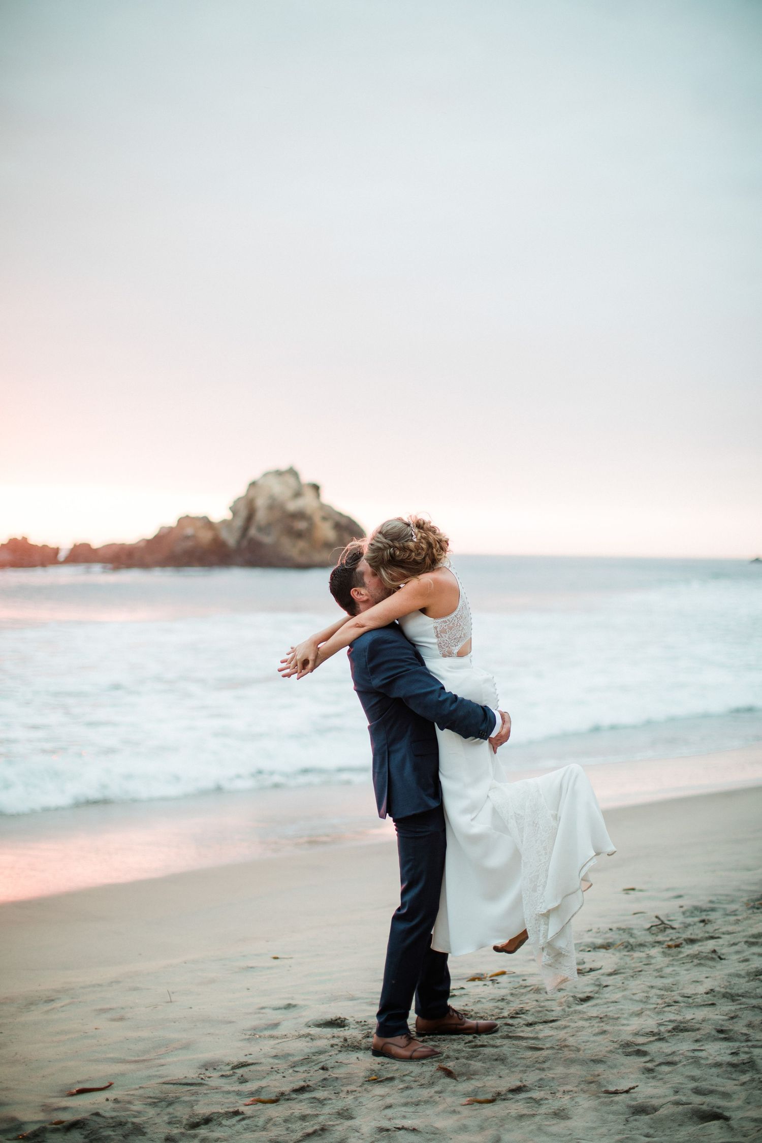 A romantic couple embraces on a sandy beach at sunset with ocean waves and rocky cliffs in the background.