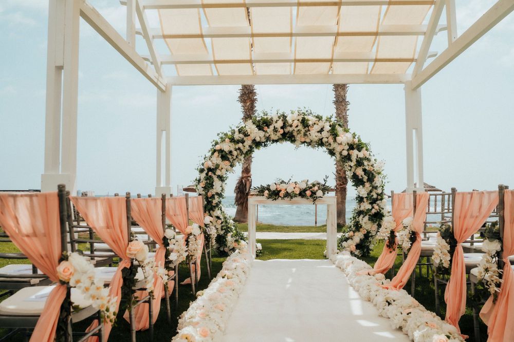 Elegant outdoor wedding ceremony setup with floral arch and coral chair sashes overlooking the ocean.