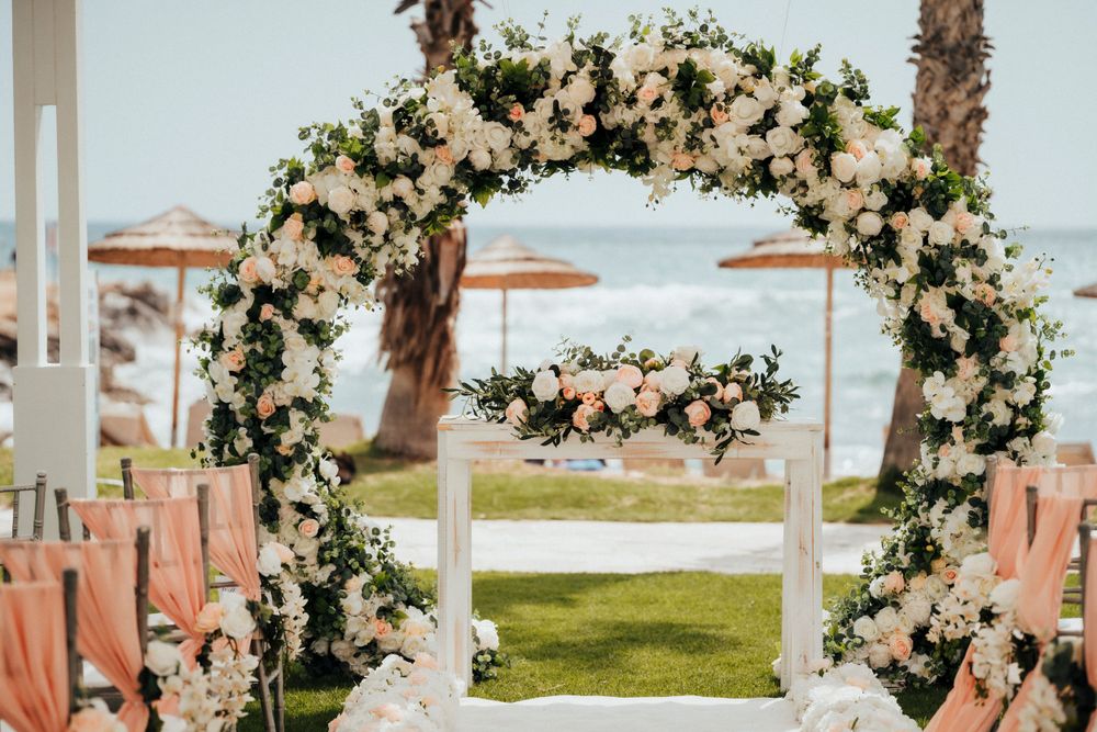 Elegant floral wedding arch with white and blush flowers overlooking the ocean and beach umbrellas.