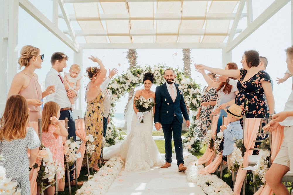 Sunlit wedding ceremony under white canopy with floral arch and guests standing along aisle as couple exits.