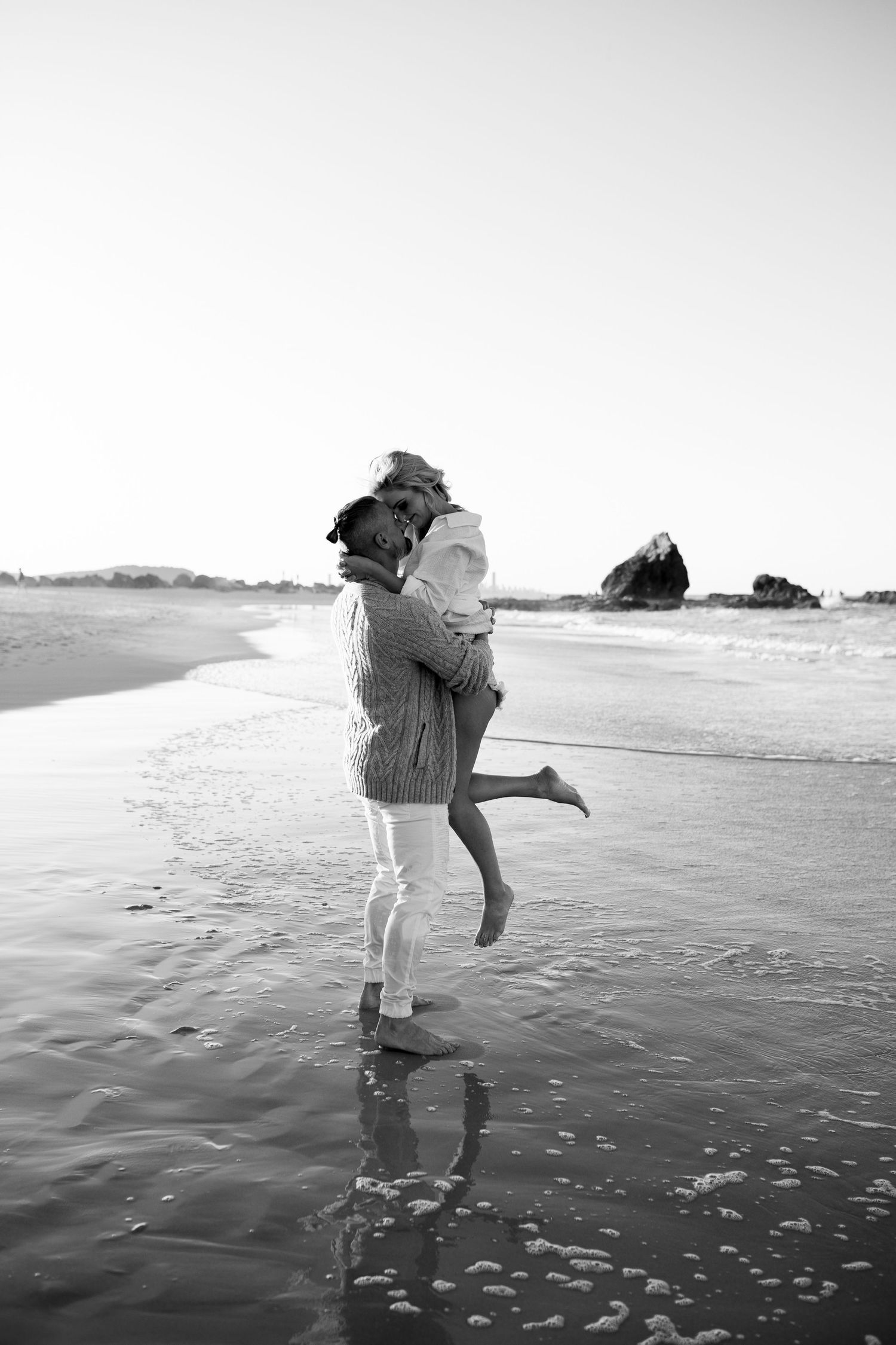 A romantic black and white photo of two people embracing on a misty beach with ocean waves and rocky cliffs in background.