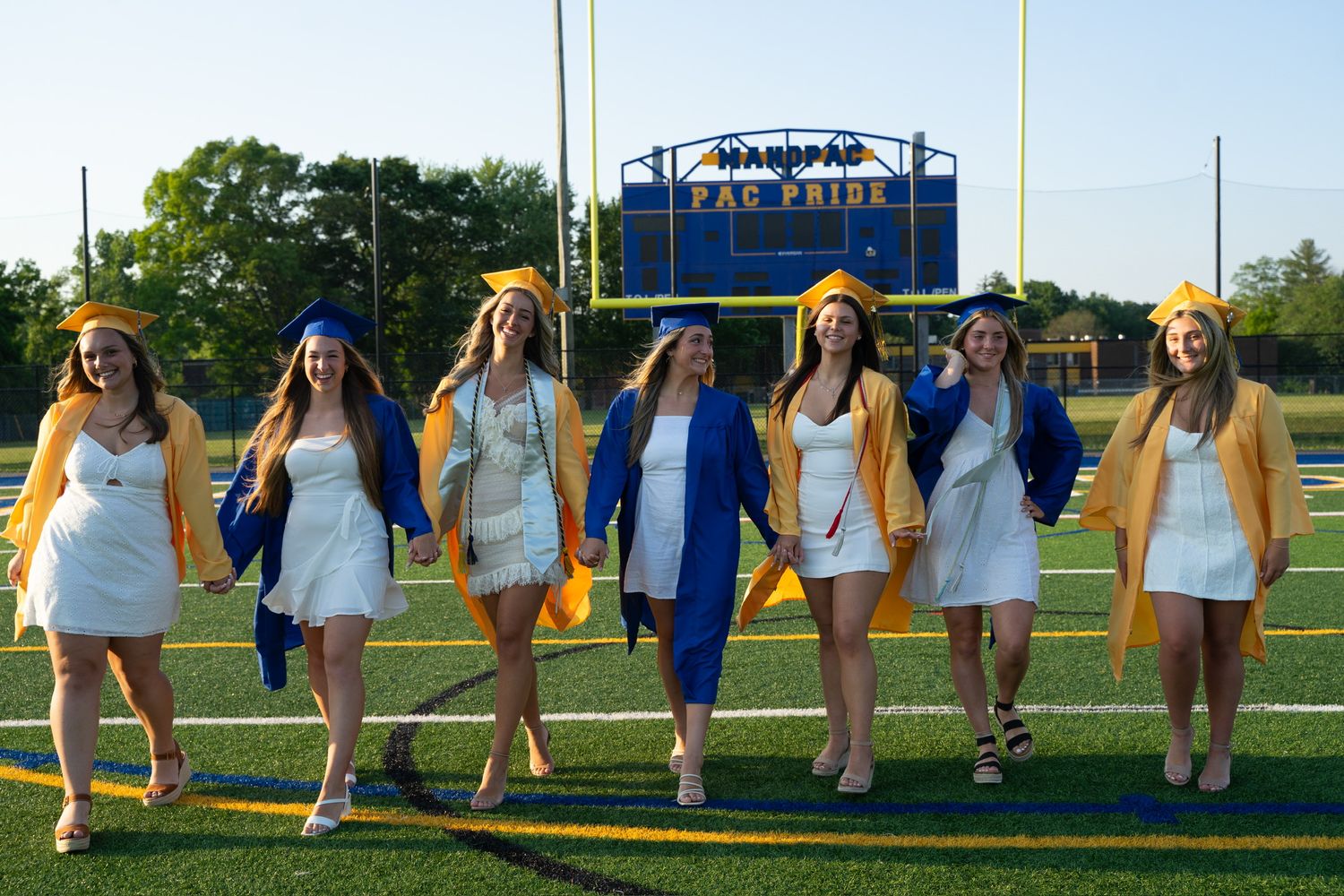 Group of graduates in white dresses and blue robes posing on a sports field.