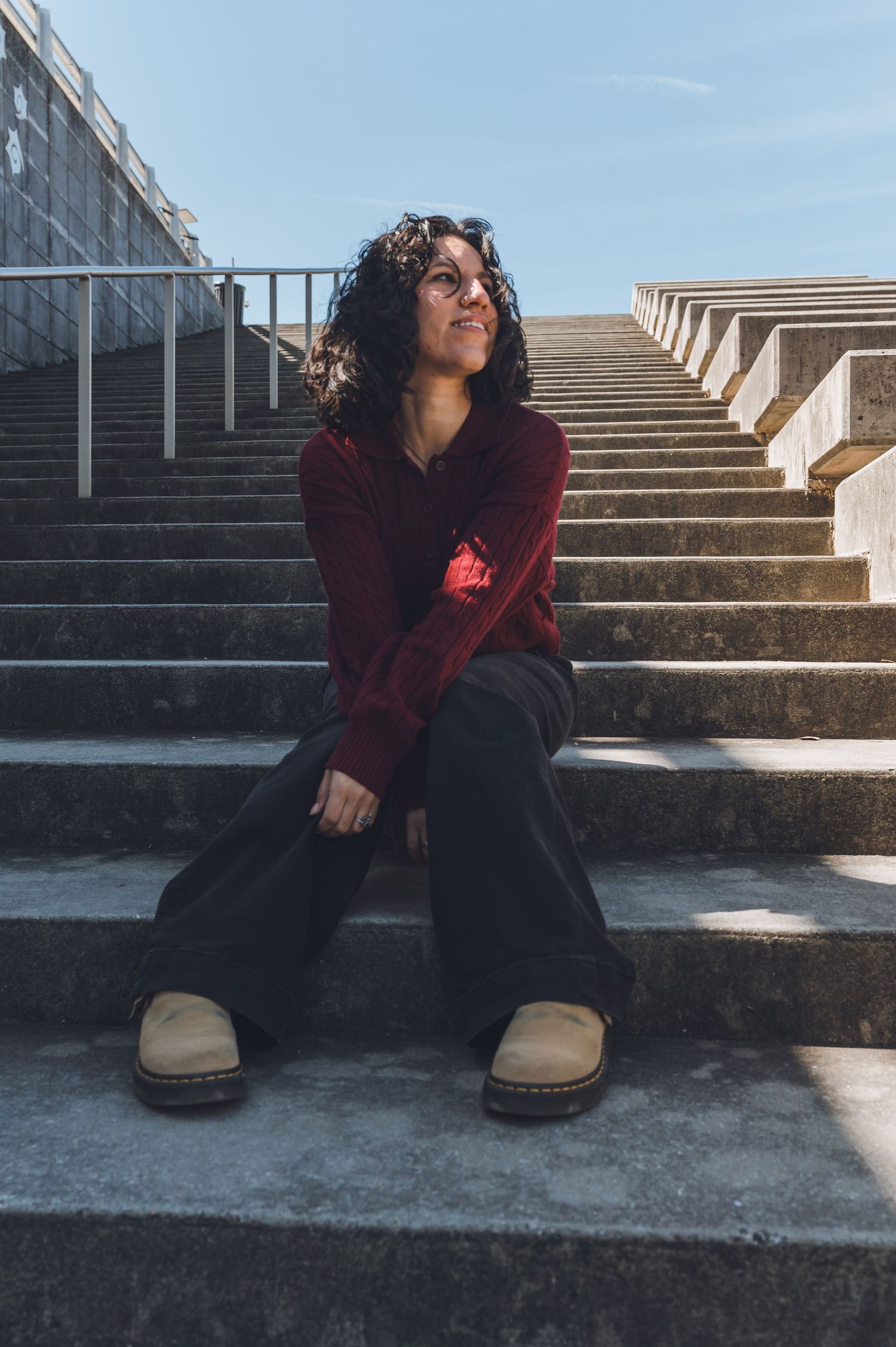 A person in a burgundy sweater sits on concrete steps in dramatic sunlight casting long shadows.