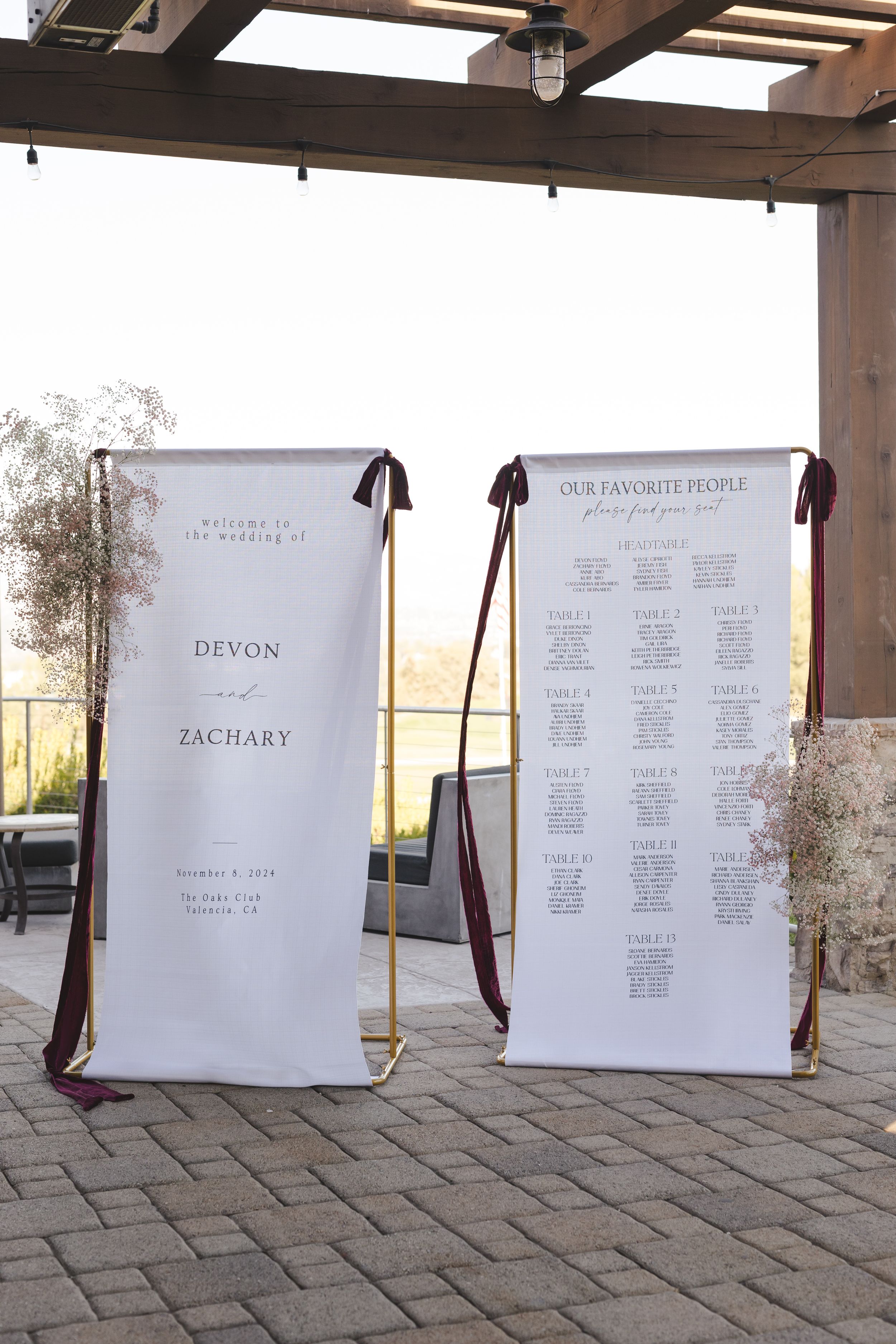Two white wedding seating charts displayed on hanging scrolls under a wooden pergola on a stone patio.