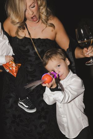 Young child in white shirt enjoying a lollipop at formal event while adult holds wine glass.