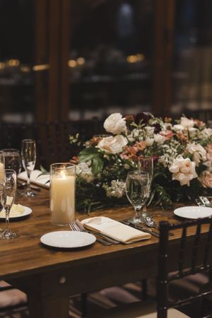 Elegant wedding table setting with white roses, candles, and glassware on a dark wooden table.