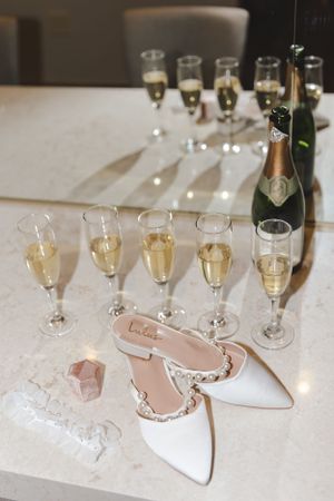 Elegant white bridal shoes next to champagne flutes and bottle on white tablecloth for wedding celebration.