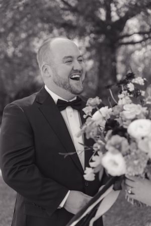 A joyful groom laughs while reading in a black tuxedo and bowtie at an outdoor wedding ceremony.