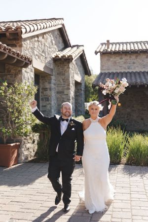 Joyful newlyweds celebrating and jumping with raised arms outside Mediterranean-style stone villa on sunny day.