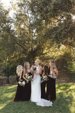 Bridal party in burgundy gowns and white wedding dress pose together on a lawn beneath sunlit trees.