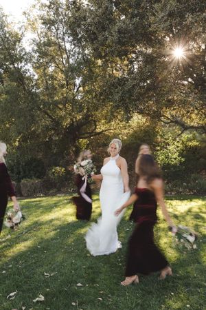 Sun streams through trees at an outdoor wedding ceremony as figures gather on a lush green lawn in late afternoon light.