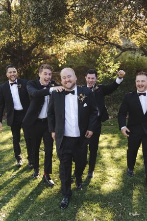 Groomsmen in black tuxedos pose playfully on a sunny lawn during wedding photos.