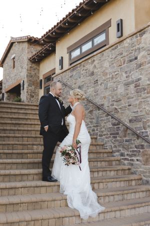 A bride and groom share an intimate moment on stone steps outside a rustic building with a beautiful bouquet.