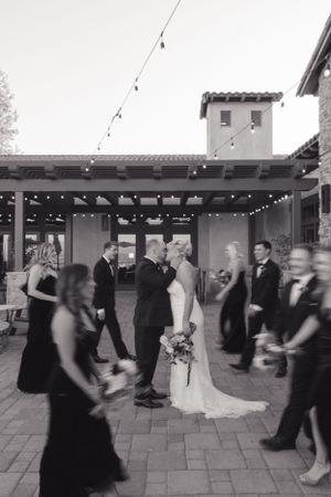 A black and white wedding reception photo showing guests dancing under string lights on an outdoor patio.