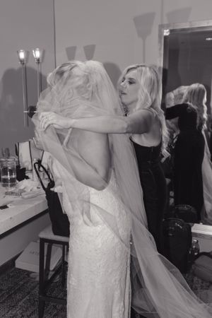 A tender embrace between a bride in a white lace dress with veil and another person in black at a wedding venue.