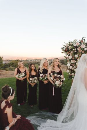Outdoor wedding ceremony setup with floral arch and bridal party in burgundy dresses at sunset.