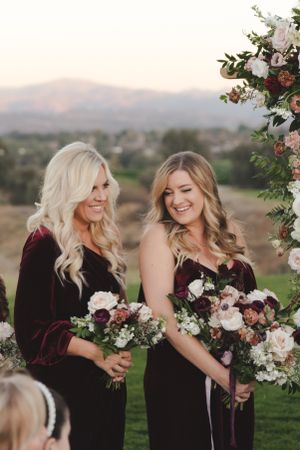 Two people wearing burgundy velvet dresses hold matching floral bouquets at an outdoor venue with mountains in background.