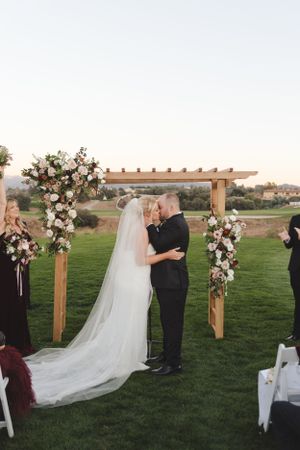 Outdoor wedding ceremony under a wooden arch decorated with pink and white flowers.