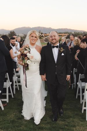 Newlyweds walk down aisle between white chairs at outdoor mountain venue.