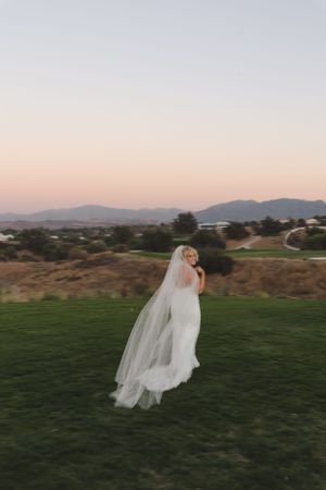 A bride in a flowing white dress and veil walks on a grassy hill during sunset with mountains in the background.