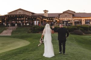 Couple walking away on golf course with Spanish-style venue and string lights in background.