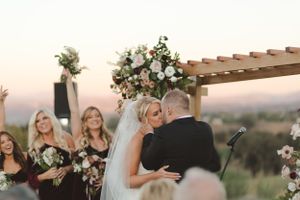 A couple shares their first kiss during an outdoor sunset wedding ceremony under a wooden pergola with floral decorations.