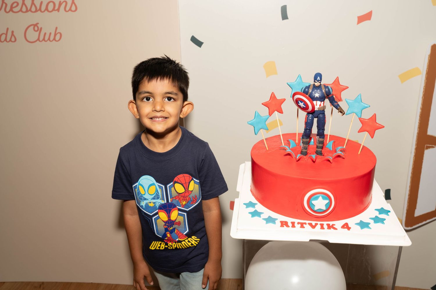 Young boy in superhero shirt standing next to a red birthday cake with decorative topper.