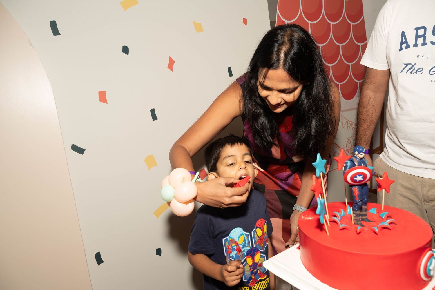 A child blows out birthday candles on a red cake while an adult helps during a celebration party.