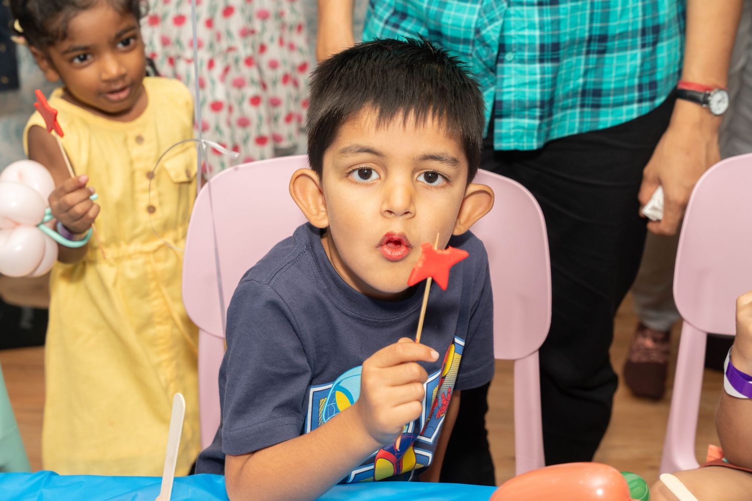 A young child in a navy sweater blows bubbles at a party with colorful decorations.