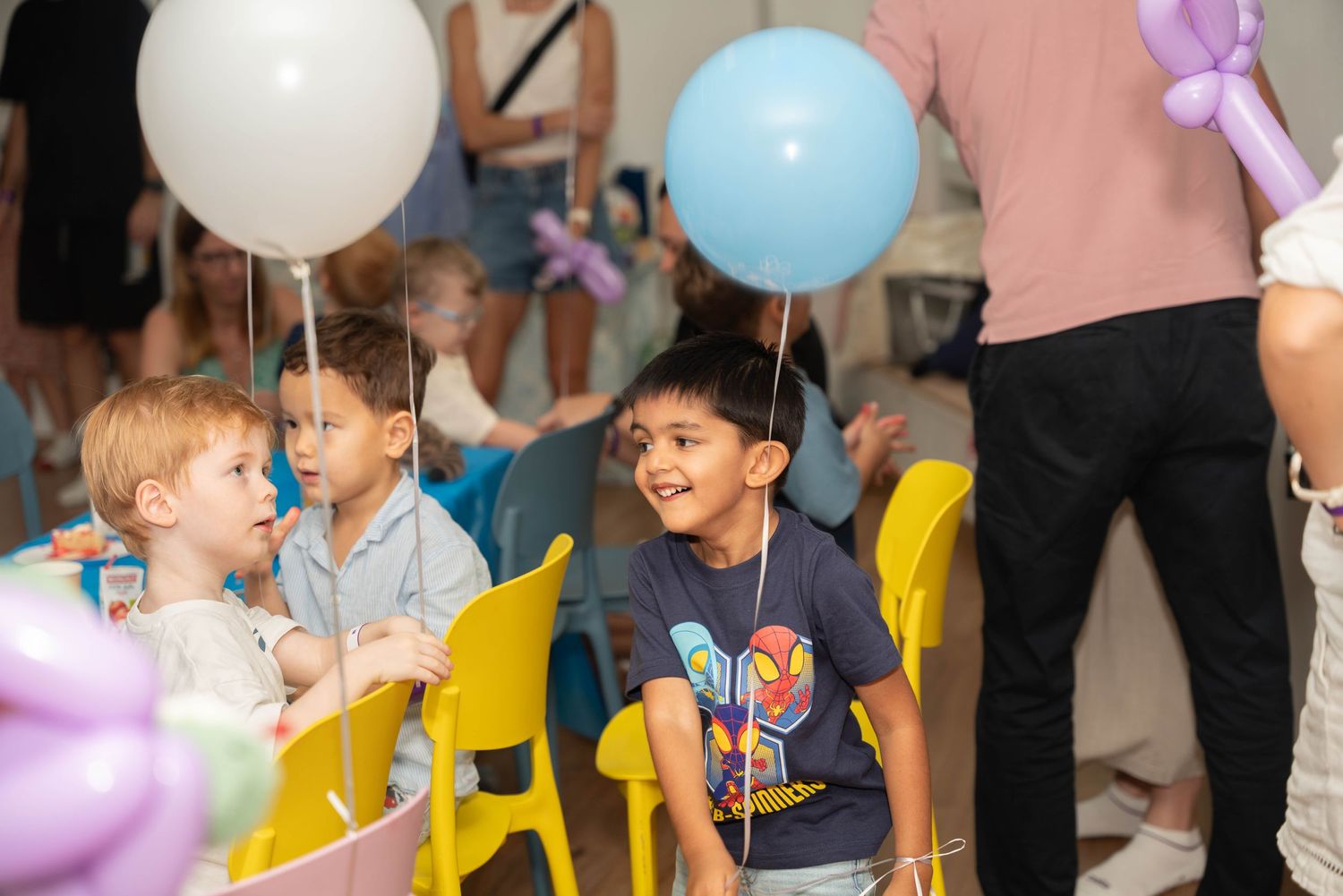 Children celebrate at a party with balloons and yellow chairs in a decorated room.