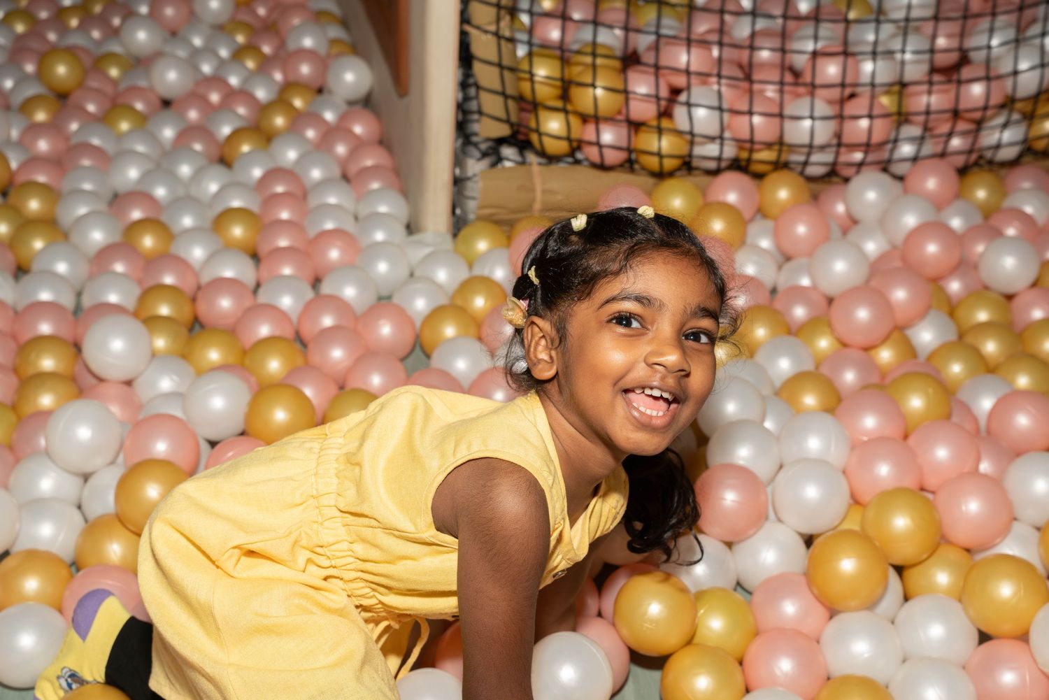 Young girl in yellow dress smiling while playing in ball pit with pink and gold balls.