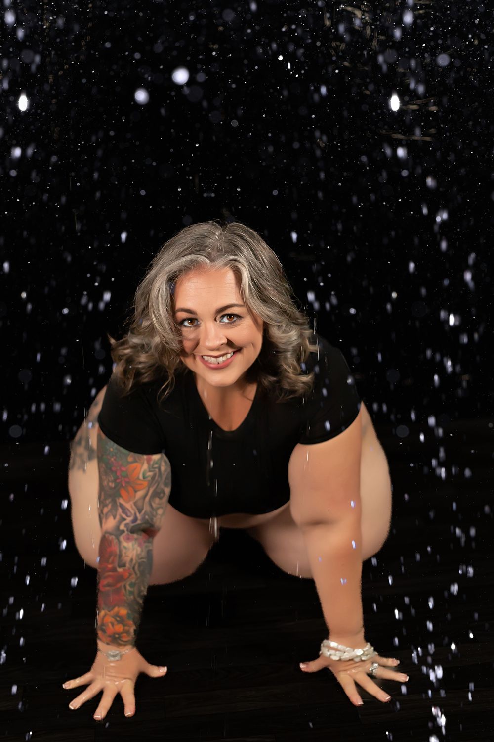 Smiling woman with gray hair and sleeve tattoo posing on black background with falling snow effect.