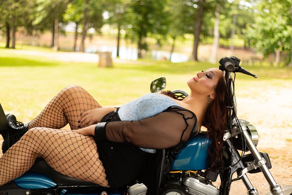 Woman in fishnet stockings posing on a classic motorcycle outdoors in a sunny park setting.
