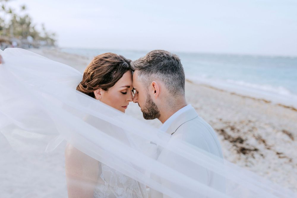 A romantic beach wedding photo featuring a couple embracing with a flowing white veil blowing in the ocean breeze.