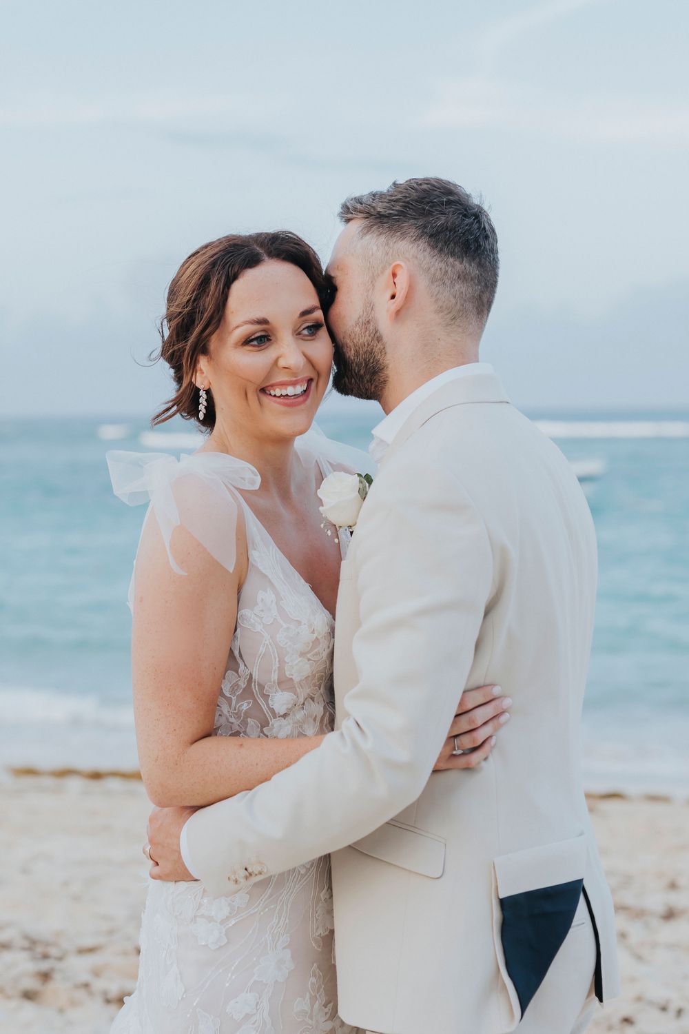 Couple in wedding attire embraces on a scenic beach with turquoise ocean waters and cloudy sky in background.