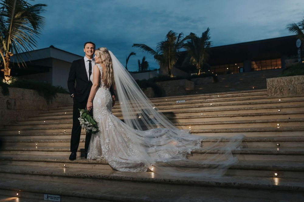 A couple stands on illuminated stone steps at dusk with a flowing wedding veil and palm trees in the background.