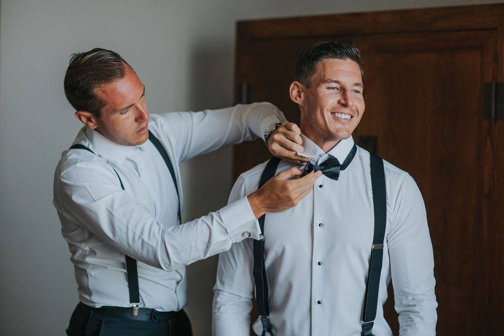 Two people in formalwear adjusting a bowtie during wedding preparations.