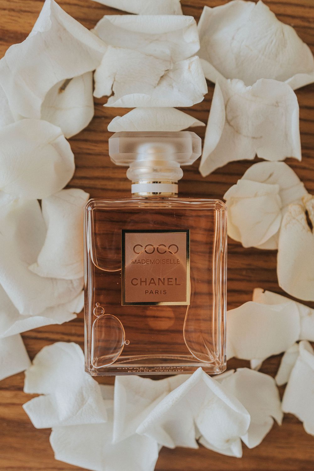A bottle of Coco Chanel perfume surrounded by white rose petals on a wooden surface.