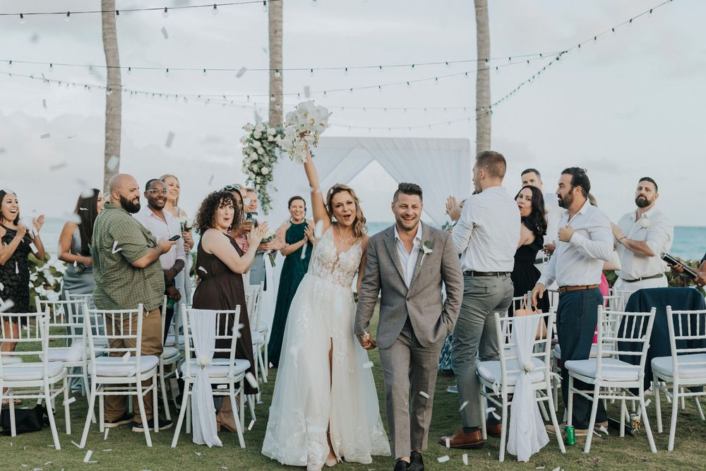Newlyweds celebrate with guests at an outdoor beach wedding reception with string lights and white chiavari chairs.
