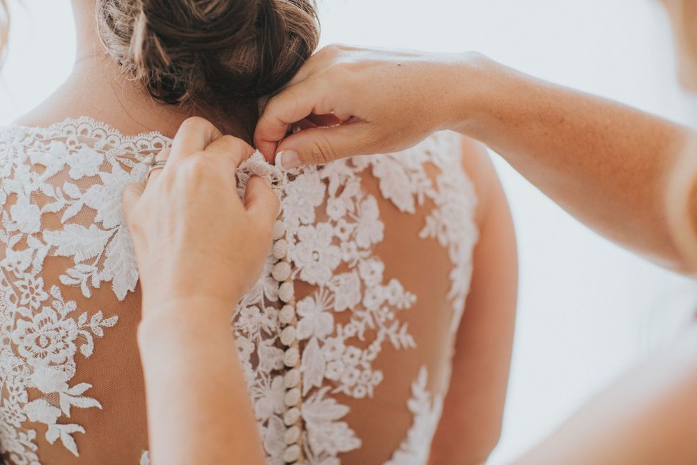 Delicate white lace detail being fastened on the back of an elegant wedding dress with floral patterns.