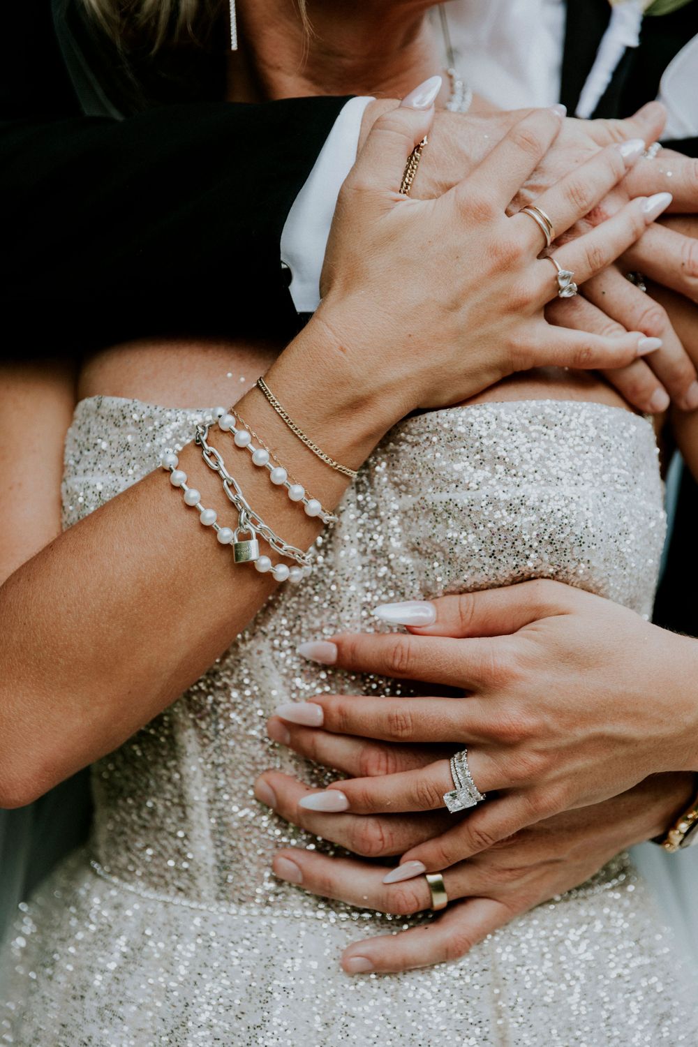 Close-up of sparkly wedding dress and multiple hands showing rings and bracelets.