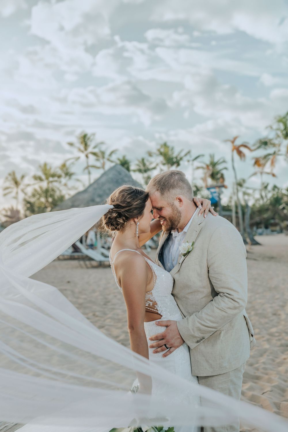 A romantic beach wedding scene with flowing veil and palm trees in the background as couple embraces on tropical sand.