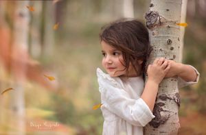 A child in a white dress hugs a birch tree trunk while autumn leaves fall in a dreamy outdoor setting.