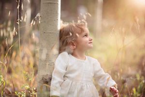 A child in a white dress stands near a birch tree in soft golden sunlight with wildflowers in the background.