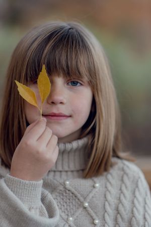 Someone in a cozy white sweater holds a yellow autumn leaf up to their face in a gentle portrait.