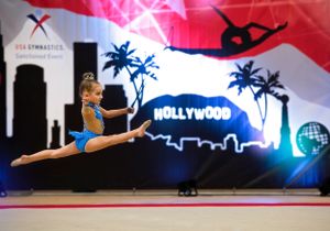 A gymnast performs a dramatic split leap against a colorful Hollywood-themed backdrop with palm trees and cityscape silhouettes.